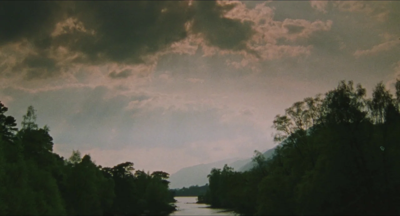 Dog Soldiers (2002) movie still: establishing — A wide shot displays a narrow river flowing between dense tree-lined banks. Distant mountains fade…