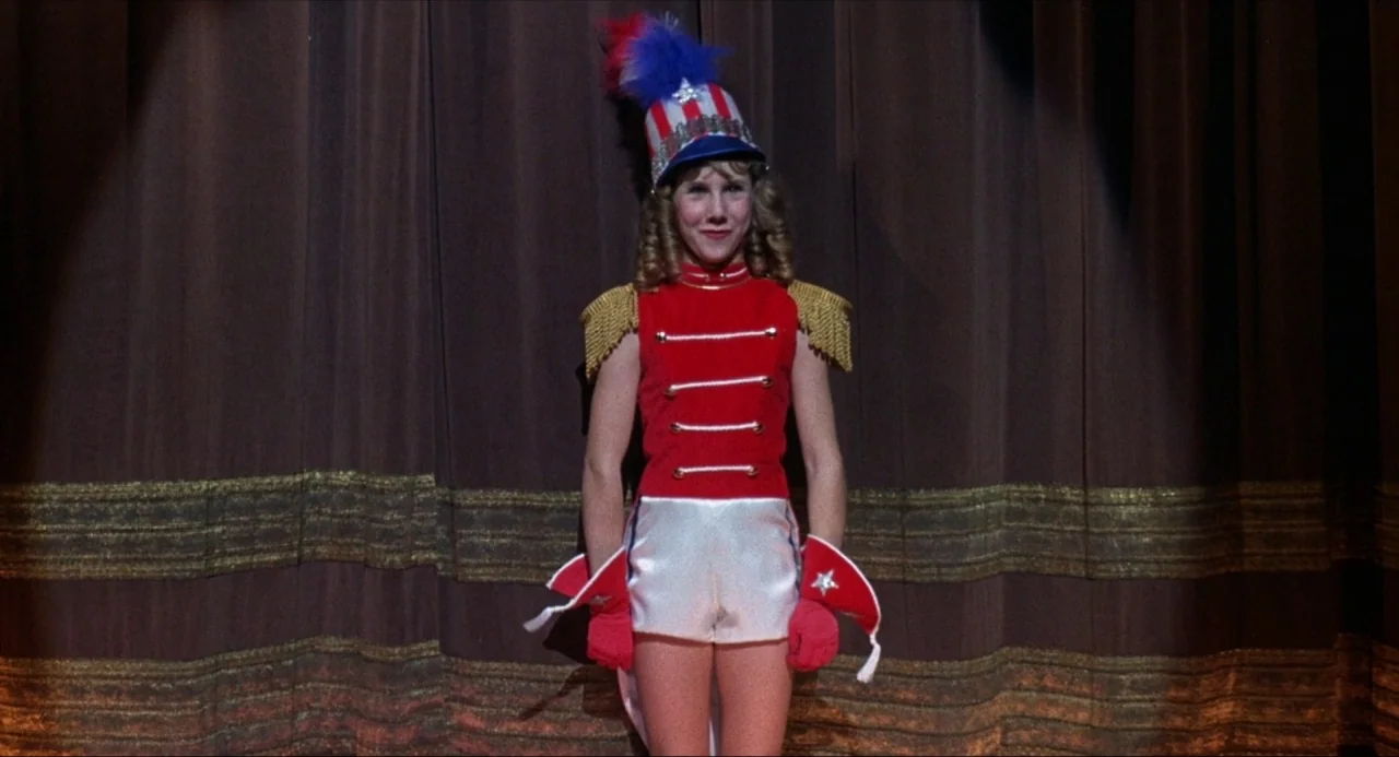 What’s the Matter with Helen? (1971) movie still: fullbody — A person stands in a parade uniform on a dark performance area. The background features…