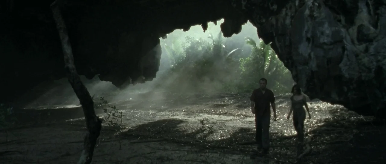 Vinyan (2008) movie still: wide — A man and a woman emerge from a large, dark rock cave into bright daylight. Lush tropical palms are visible in the…
