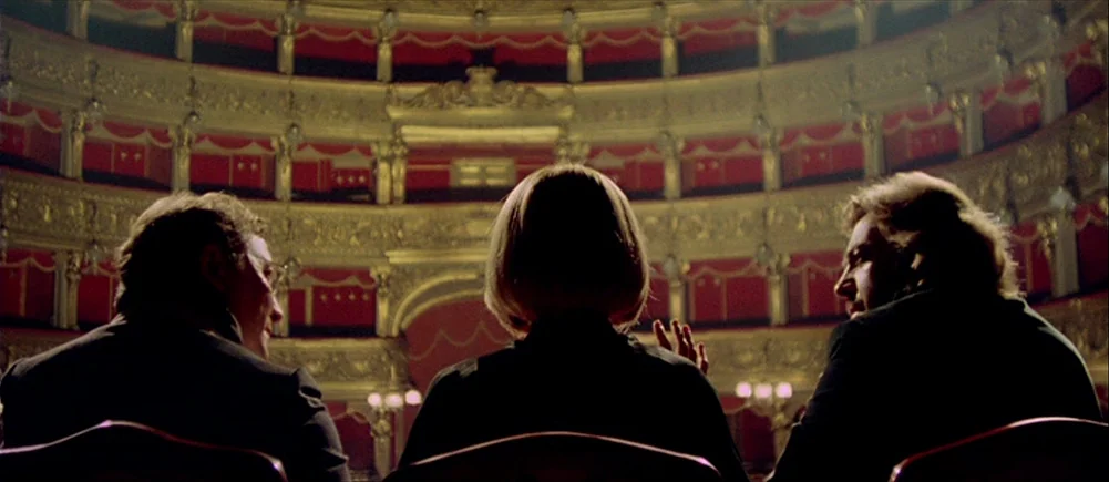 Deep Red (1975) movie still: medium — A historic opera house with ornate gold-trimmed balcony seating and deep red velvet walls. Three people sit in…