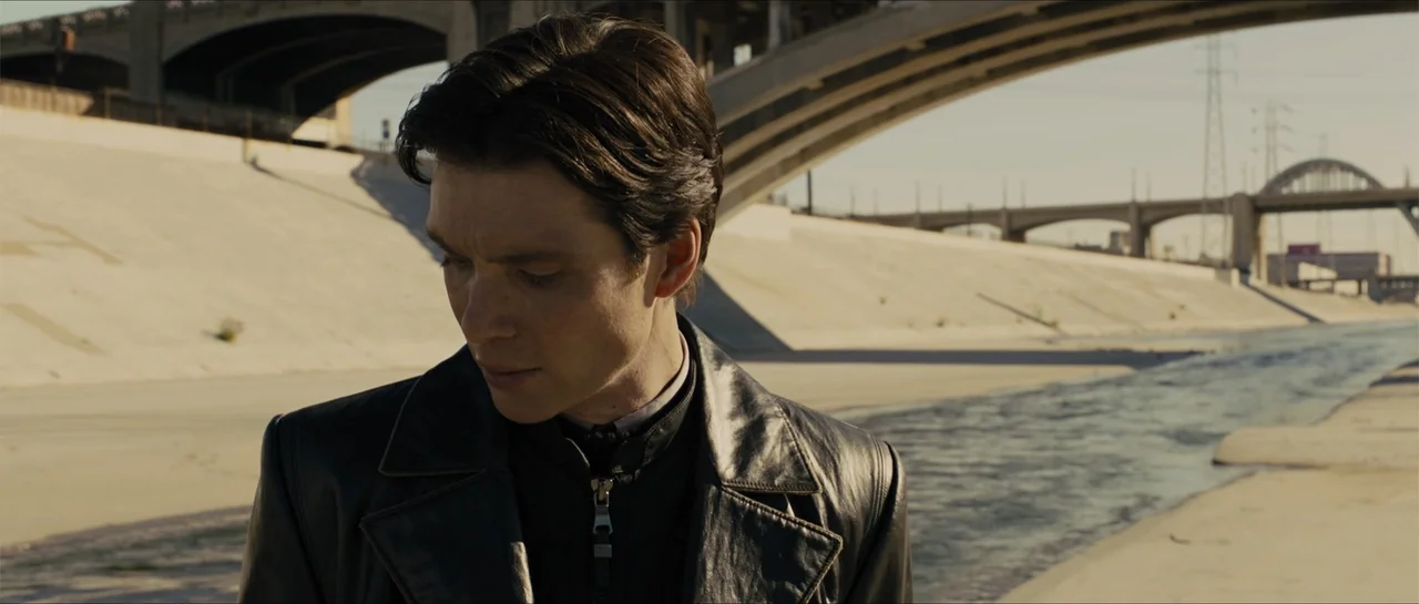In Time (2011) movie still: medium — A man stands in a vast concrete flood control channel under a concrete bridge. He gazes downward while…