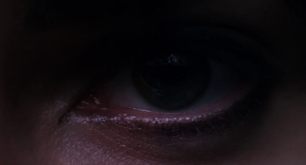 Under The Skin (2013) movie still: closeup — An extreme close-up of a human eye in near darkness. The eye is partially obscured by shadows while the…