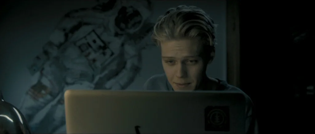 Lasting (Nieulotne) (2013) movie still: medium — A dark room features an astronaut poster hanging on the wall behind the subject. A young man sits…