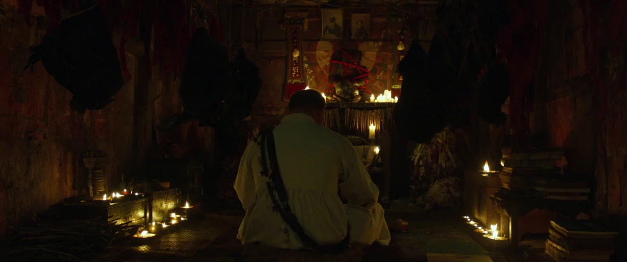 The Wailing (2016) movie still: medium — A dimly lit room filled with religious artifacts and candles burning in small bowls. An elderly man sits on…