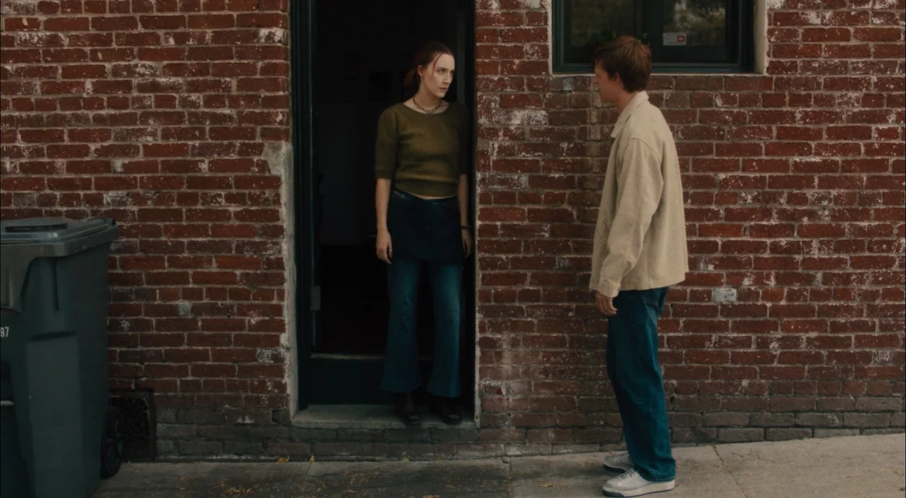 Lady Bird (2017) movie still: fullbody — A red brick wall forms the backdrop to an exterior doorway on a sidewalk. A young woman stands in the…