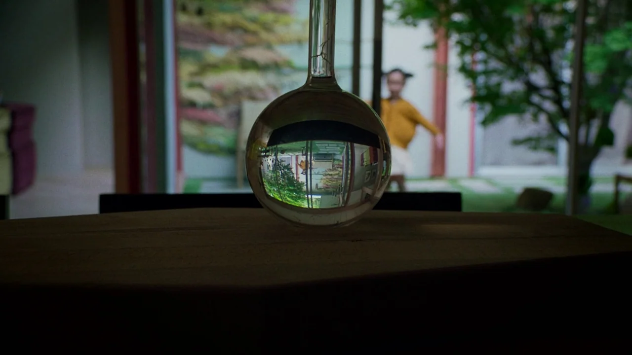 After Yang (2021) movie still: closeup — A dark wooden surface sits in the foreground with a clear glass bulb resting atop. Inside the reflection of…