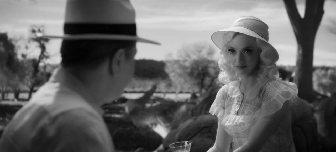 Mank (2020) movie still: medium — An outdoor park setting with trees visible in the soft-focused background area. A woman wearing a hat looks toward…