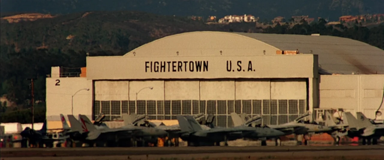 Top Gun (1986) movie still: establishing — A large white military hangar is labeled Fightertown U.S.A. in black capital letters. Several grey…