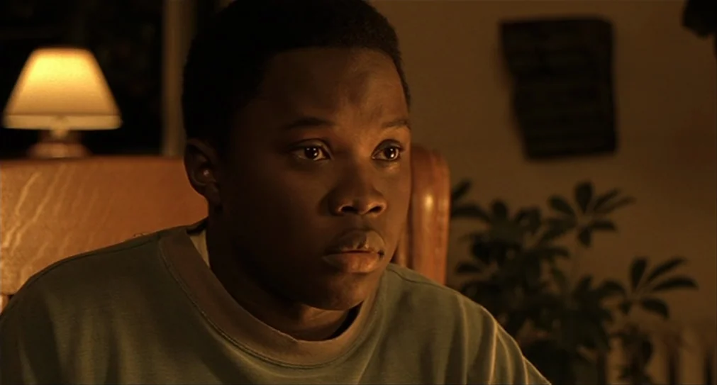 The Hurricane (1999) movie still: closeup — A dimly lit room contains a table lamp, a houseplant, and wooden furniture. A teenager looks toward an…