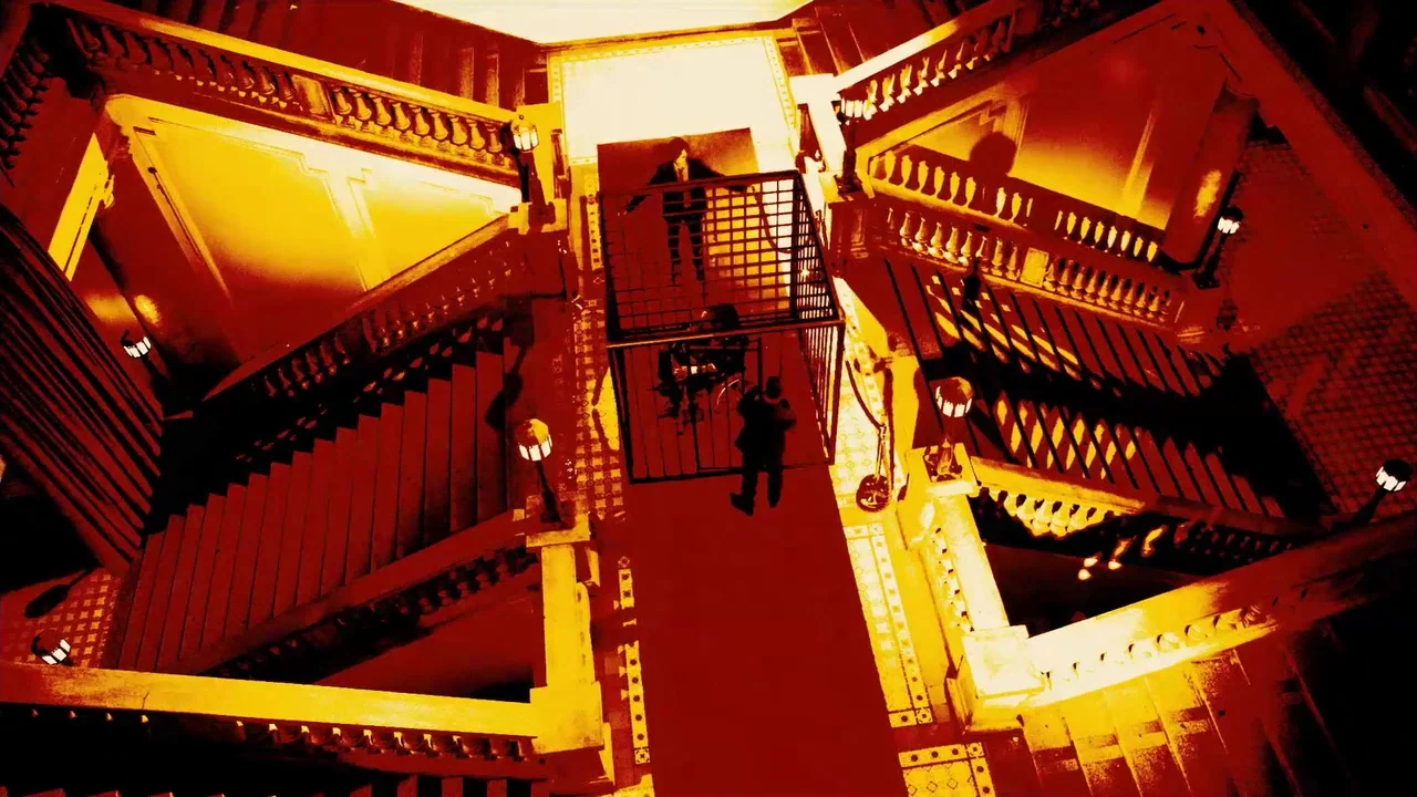 The ABCs of Death (2012) movie still: wide — A grand staircase with ornate balustrades surrounds a central metal cage on the landing. One person…
