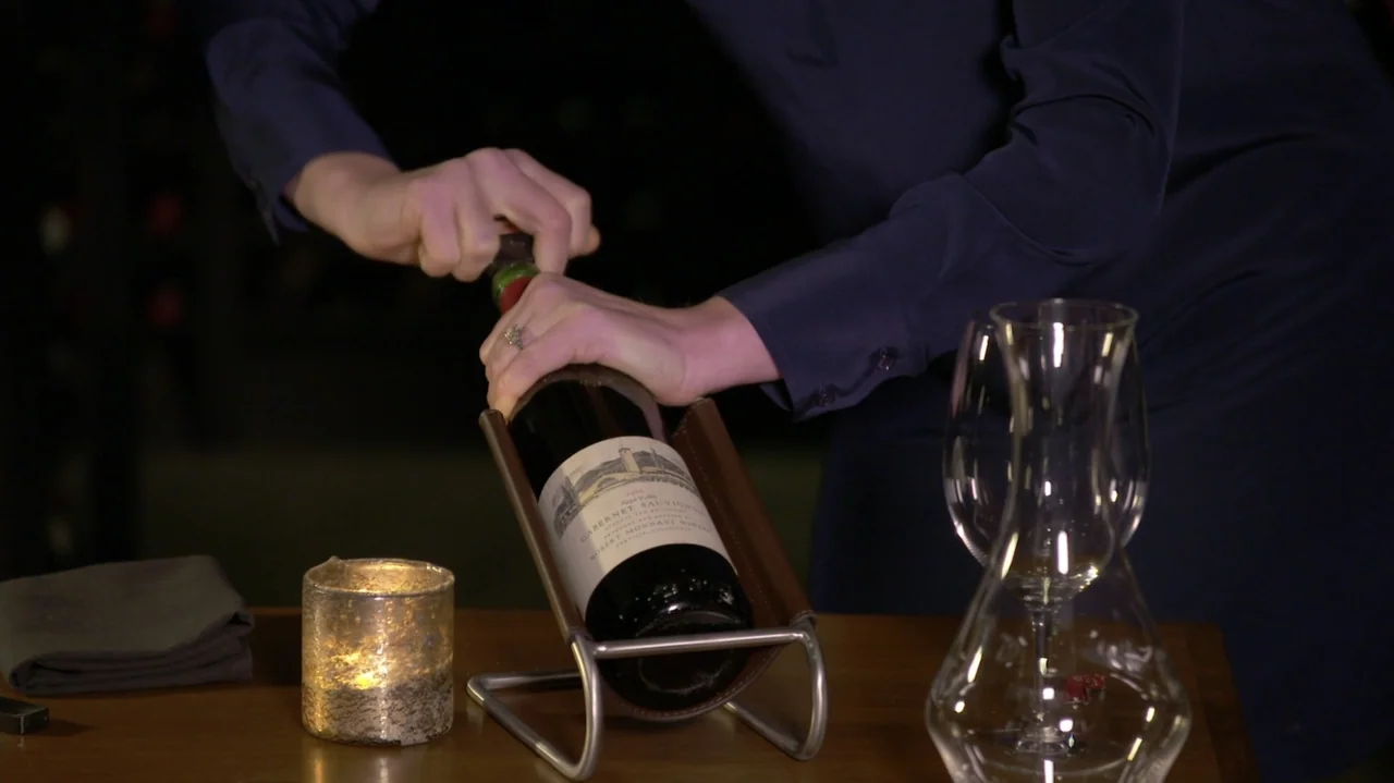 Somm: Into the Bottle (2015) movie still: medium — A dimly lit table holds a wine bottle in a leather cradle and two empty glasses. A person in a…