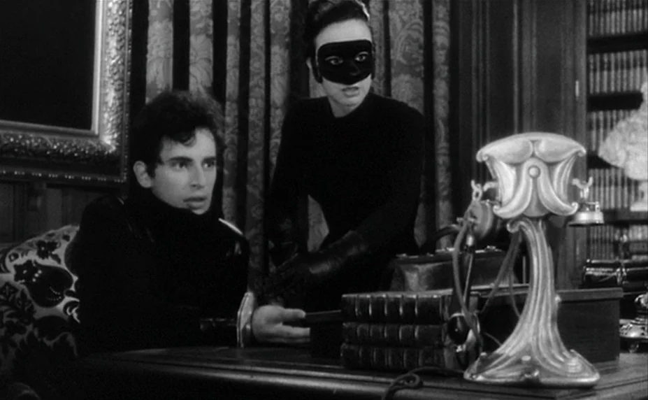 Judex (1966) movie still: medium — A dimly lit room features heavy curtains, a wooden desk, and a bookshelf. A masked woman stands beside a man…