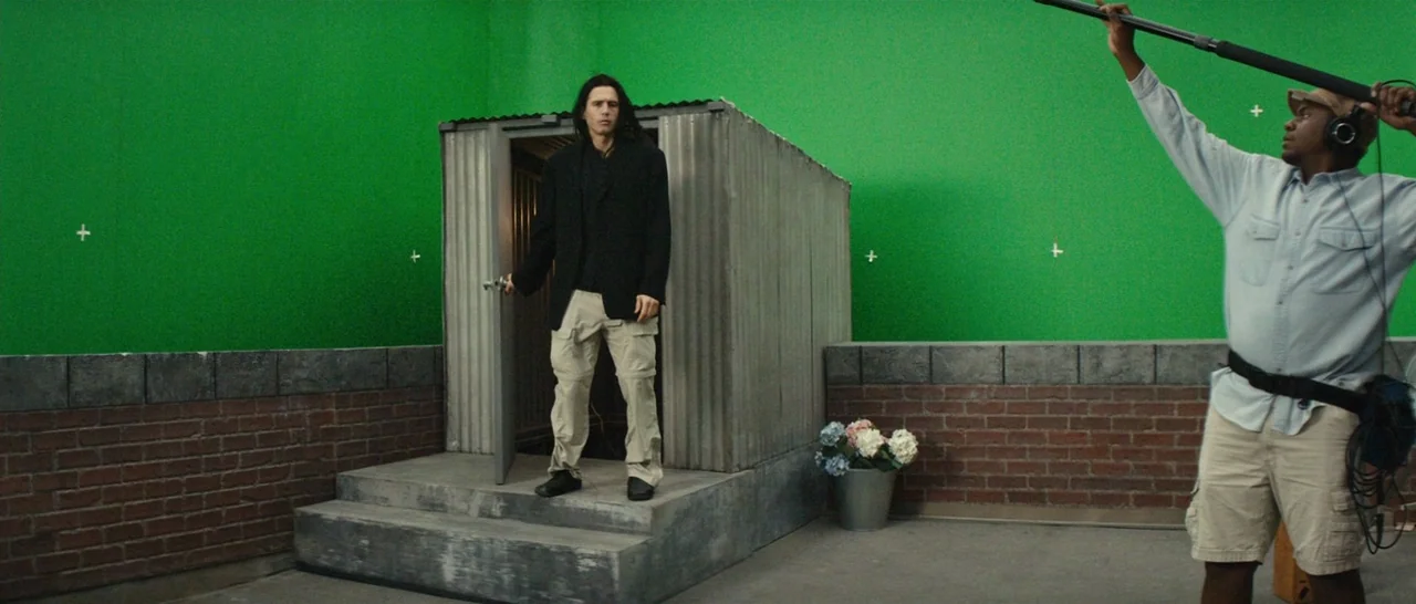 The Disaster Artist (2017) movie still: wide — A film set features a green screen wall and a corrugated metal shed structure. A man in black stands…