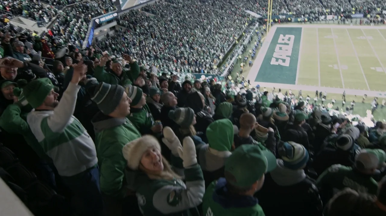 SAQUON (2025) movie still: wide — A dense crowd of football fans fills the stadium seating during a game. The spectators cheer and wave hands while…