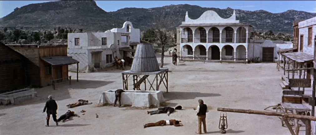 Fistful Of Dollars (1964) movie still: wide — A dusty, sun-drenched frontier town plaza is surrounded by weathered adobe and wooden buildings.…