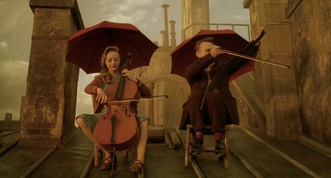 Delicatessen (1991) movie still: fullbody — A man and woman play musical instruments on a painted rooftop set. Both musicians sit on wooden stools…