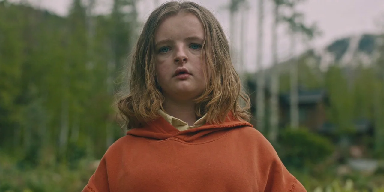 Hereditary (2018) movie still: medium — A teenage girl stands in an overgrown woodland area with birch trees in the background. She faces forward…