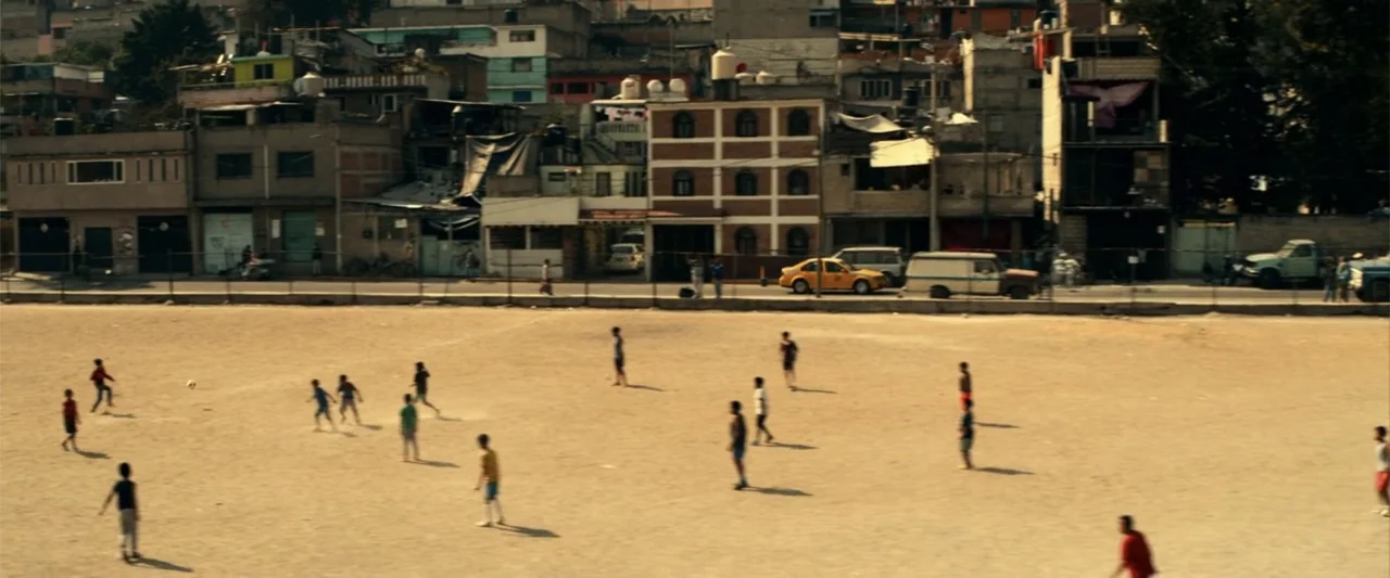 Bad Boys for Life (2020) movie still: wide — A dry, sandy dirt soccer field sits before a dense urban neighborhood of buildings. A group of children…