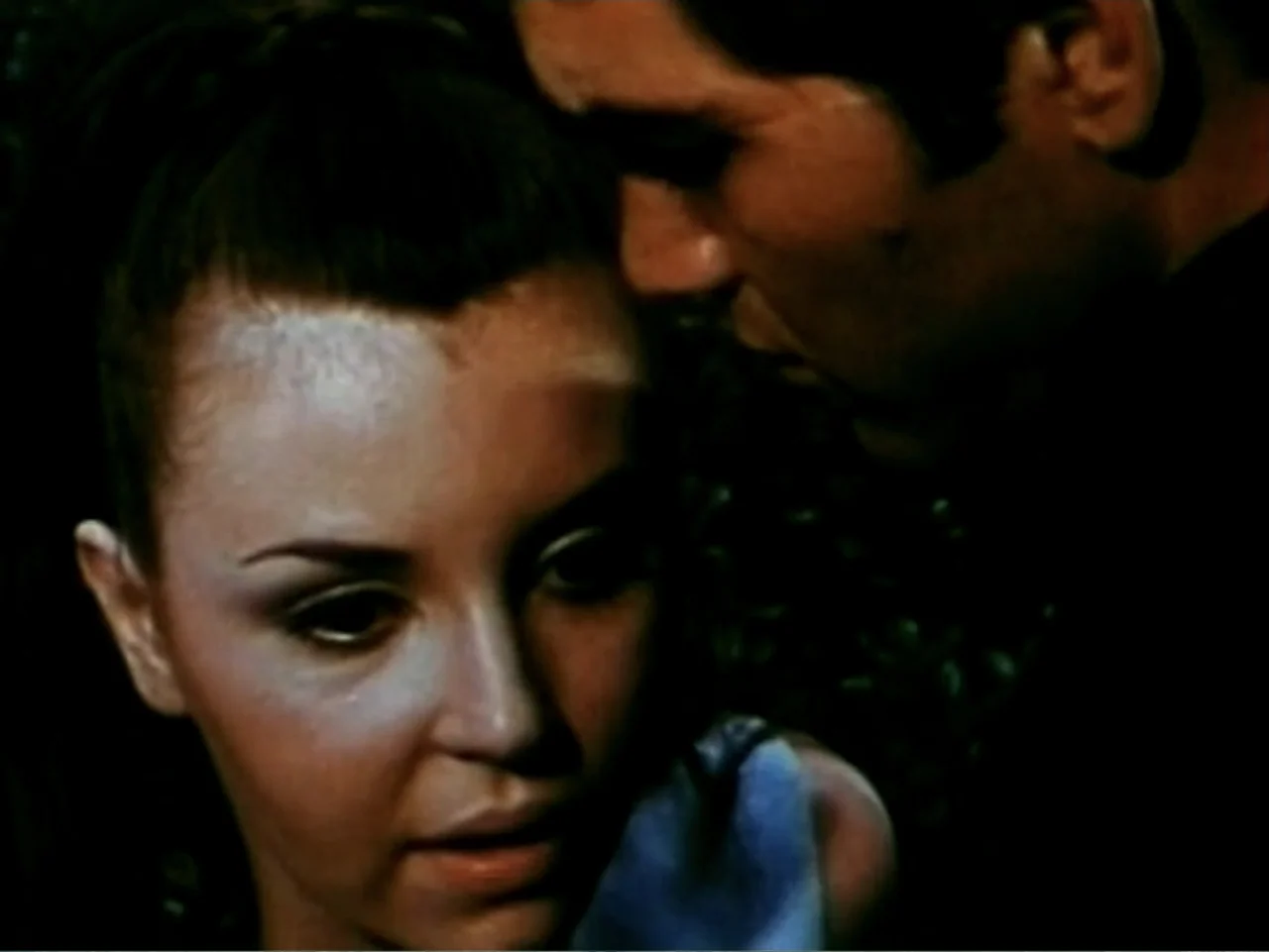 In the Year 2889 (1969) movie still: closeup — A close perspective shows a woman in a low light setting. A man standing behind her leans toward her…