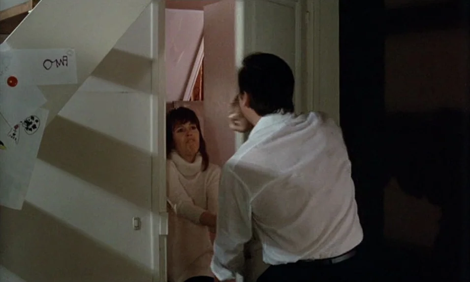 Tout Va Bien (1972) movie still: medium — A dark, narrow residential interior with white walls and drawings taped to the stairwell. A man stands in…