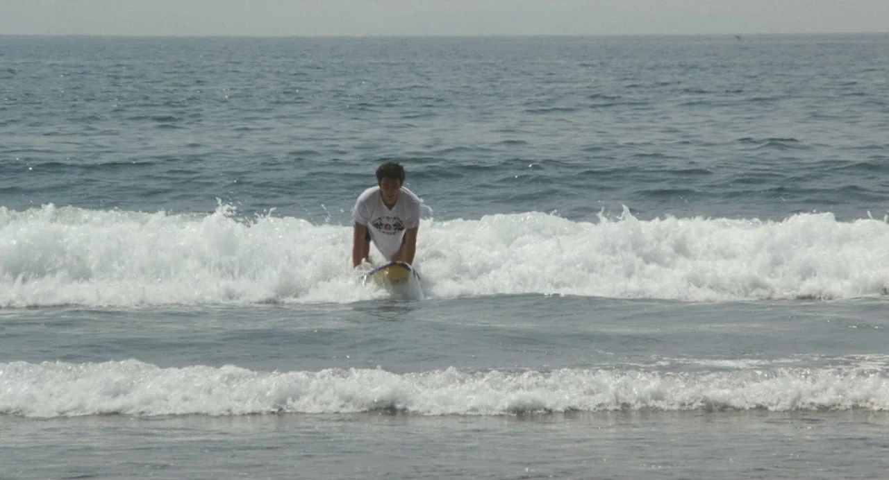 A Scene at the Sea (1991) movie still: wide — A wide view shows a surfer riding a small wave in open water. The man pushes himself up on his…