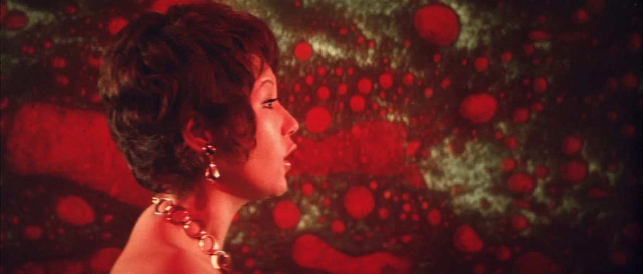 Godzilla Vs Hedorah (1971) movie still: closeup — A woman stands against a textured, liquid-like backdrop of swirling red and dark patterns. She is…