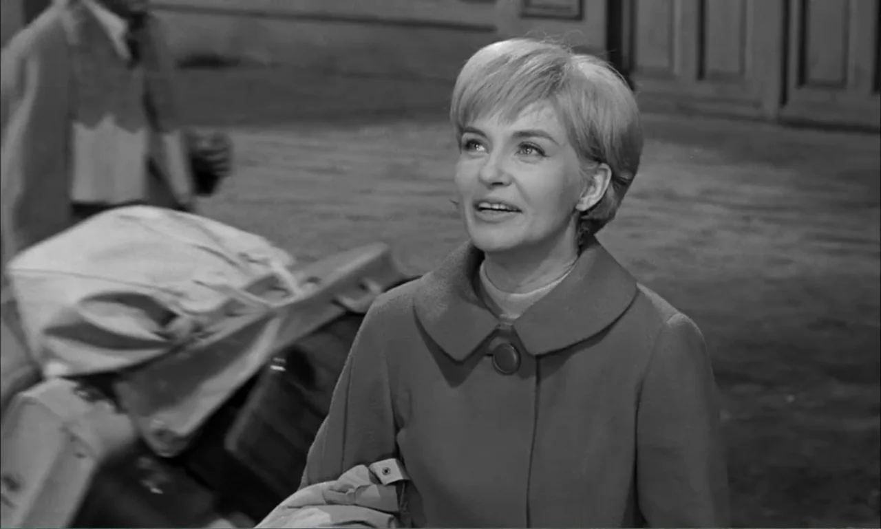 Paris Blues (1961) movie still: medium — A woman stands in a paved outdoor area with a suitcase near her. She gazes upwards into the distance while…