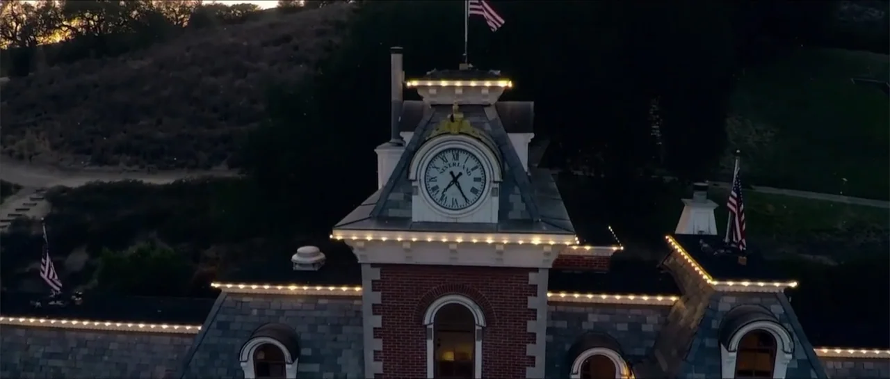 Square One (2019) movie still: establishing — A stone and brick clock tower stands at dusk with lit roofline trim. Two American flags fly from the…