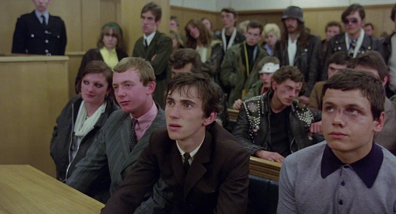 Quadrophenia (1979) movie still: medium — A crowded public gallery features rows of wood-paneled benches filled with young adults. They sit in…