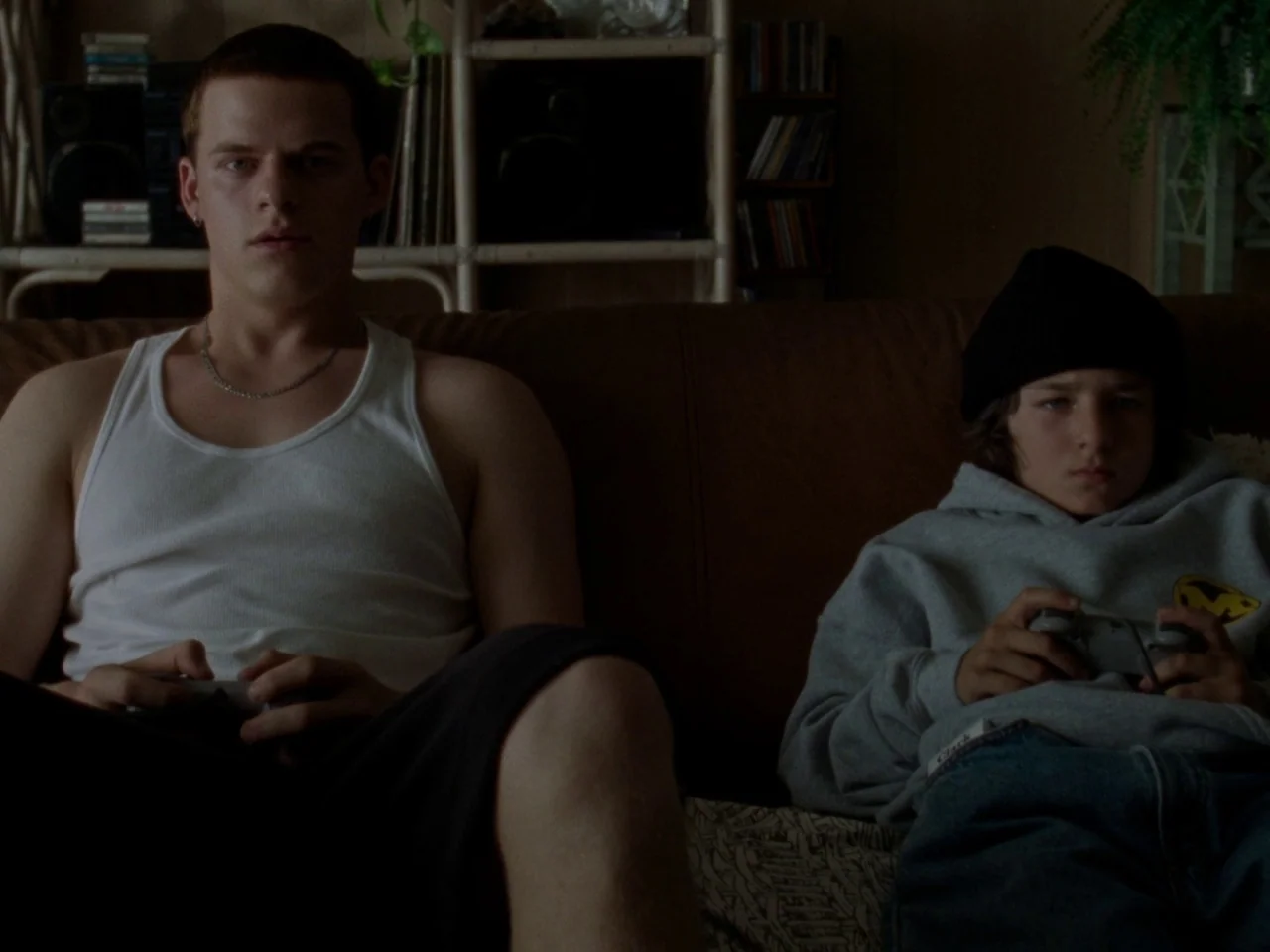 Mid90s (2018) movie still: medium — Two young men sit on a brown leather couch in a cluttered living room. They hold video game controllers while…