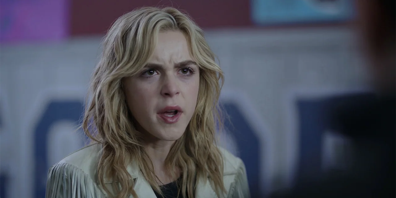 Totally Killer (2023) movie still: closeup — A school hallway features a blue wall with lockers and blurred architectural details. A blonde woman…