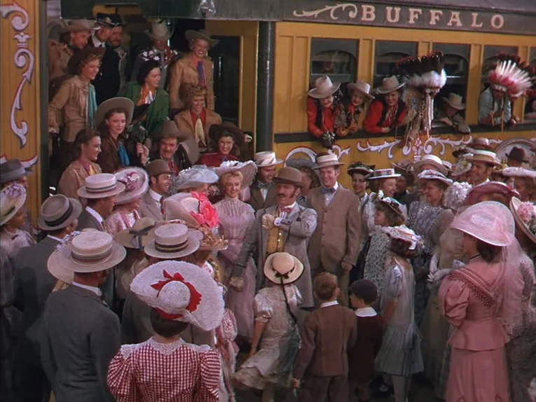Annie Get Your Gun (1950) movie still: wide — A crowd of people in period clothing gathers around a yellow Buffalo Bill train car. Individuals in…