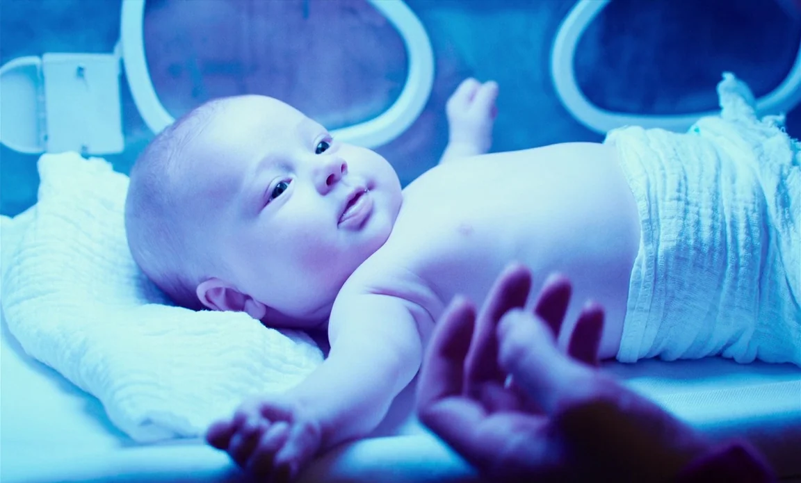 High Life (2018) movie still: closeup — A newborn baby lies under a blue phototherapy lamp inside a medical incubator enclosure. An adult hand is…
