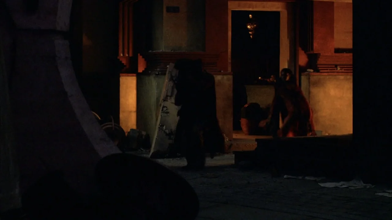 The Chronicles of Riddick (2004) movie still: medium — A dark outdoor space features an illuminated doorway set into a stone wall. Two figures move…
