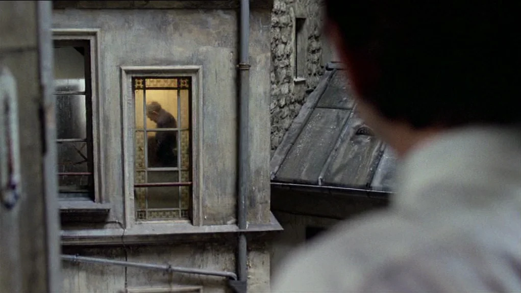 The Tenant (1976) movie still: medium — An aging grey stone apartment building shows a small window with a lit interior. A man stands inside the…