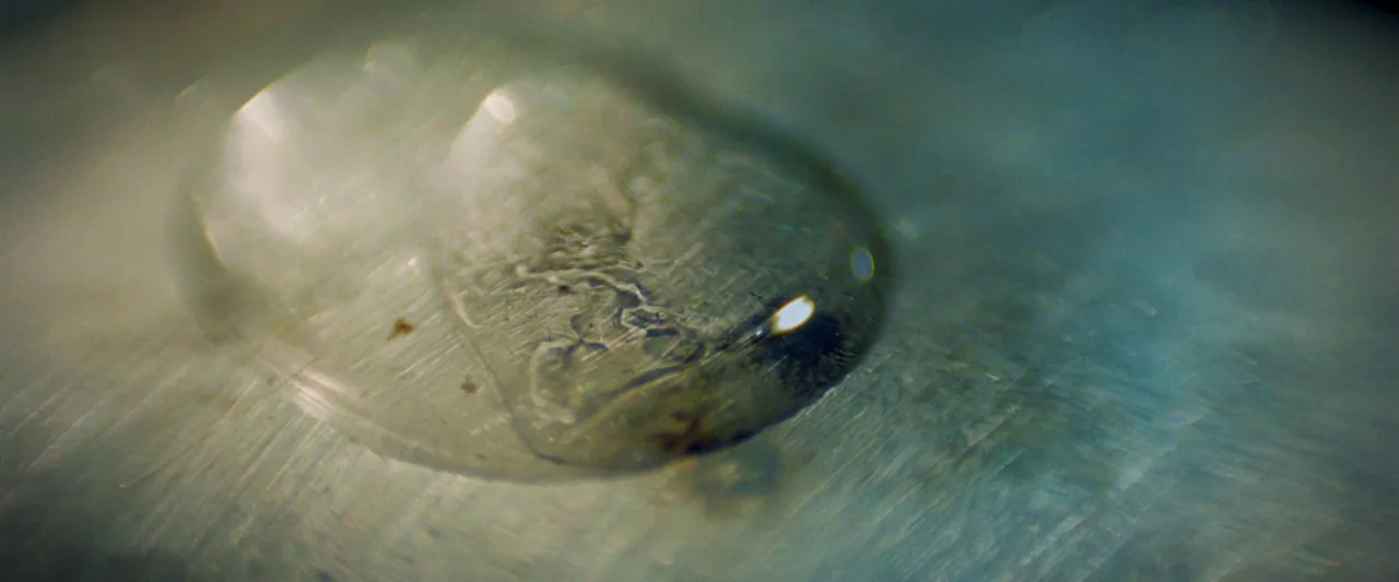 Moon Garden (2022) movie still: closeup — A translucent droplet sits on a brushed metallic surface with visible fine scratches. The liquid creates a…