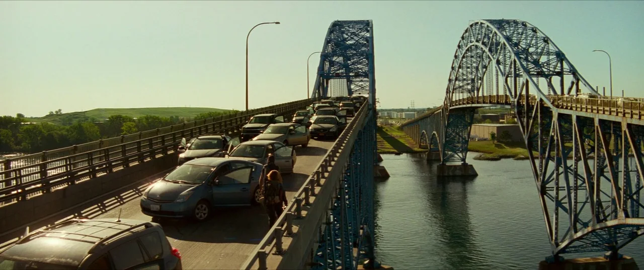 A Quiet Place Part II (2020) movie still: wide — A long blue metal truss bridge spans across a wide body of water. Abandoned cars line the lanes…