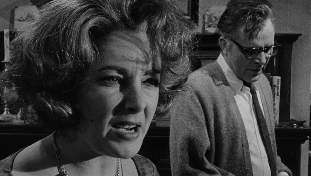 Who’s Afraid Of Virginia Woolf? (1966) movie still: medium — A dimly lit room with bookshelves and a fireplace visible in the background. A woman…