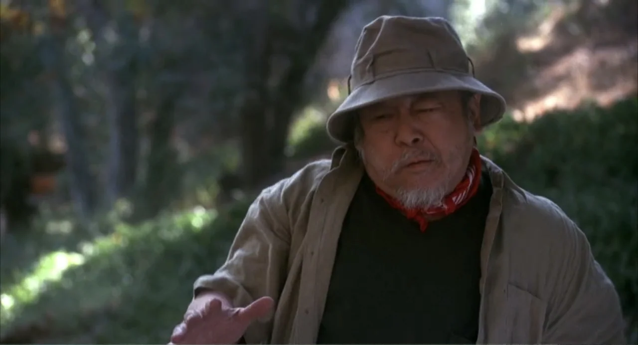 3 Ninjas (1992) movie still: medium — A forested area with dense green foliage and blurred trees in the background. An elderly man in a beige hat…