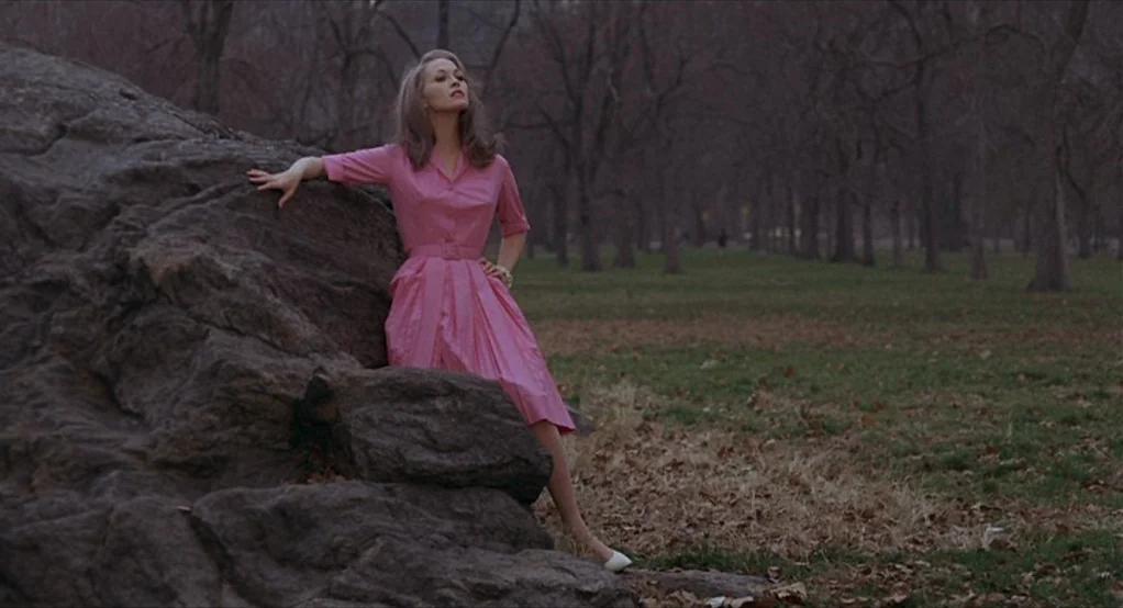Puzzle of a Downfall Child (1970) movie still: fullbody — A woman stands in a grassy park area beside a massive grey rock formation. Bare trees fill…