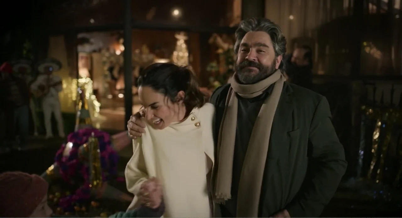 La posada (2023) movie still: medium — A nighttime outdoor gathering features a man and woman walking together outdoors. The couple smiles and…