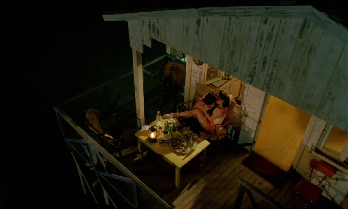 Betty Blue (1986) movie still: wide — A dimly lit wooden porch at night features a small table with a lantern. A couple sits together in chairs…