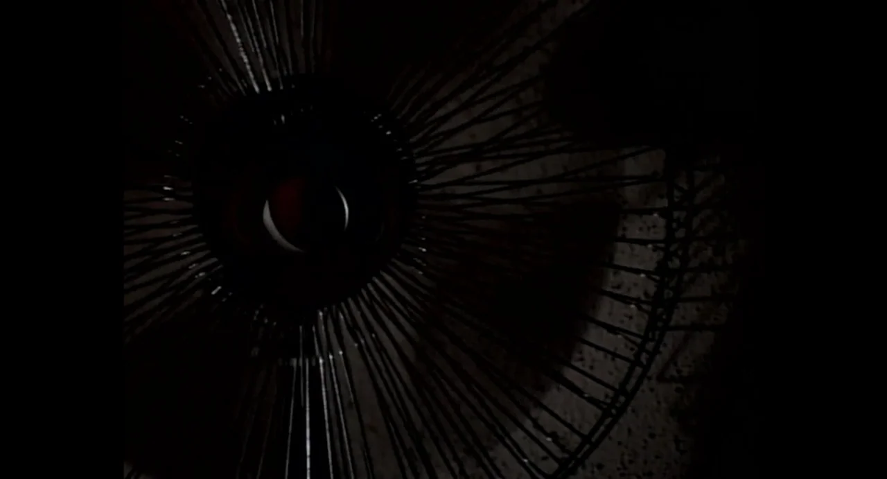 American Yakuza (1993) movie still: closeup — A close-up view shows the dark wire cage of a vintage electric fan. The central hub is dimly lit…
