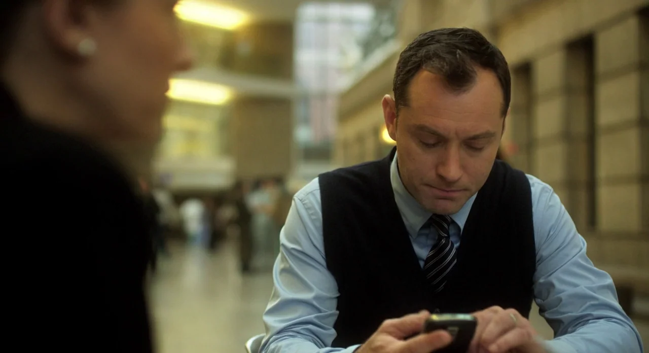 Side Effects (2013) movie still: medium — A man in a vest and tie sits at a table in a vast, stone-lined building. He checks his smartphone while a…
