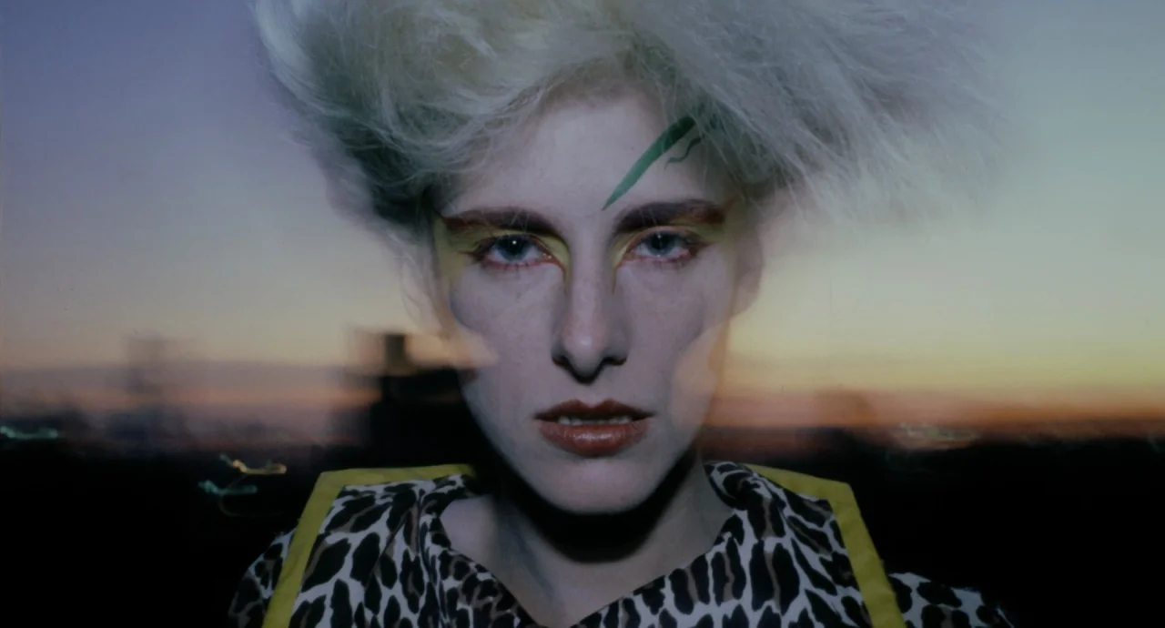 Liquid Sky (1982) movie still: closeup — A close portrait of a person with spiky blonde hair and distinct graphic makeup. The subject gazes forward…