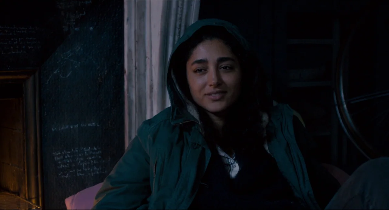 The Night Eats the World (2018) movie still: medium — A dimly lit room with a black wall covered in faint white chalk writing. A woman wearing a…