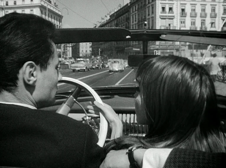 Le Petit Soldat (1963) movie still: medium — A man drives a convertible through a European city street lined with tall buildings. The passenger sits…