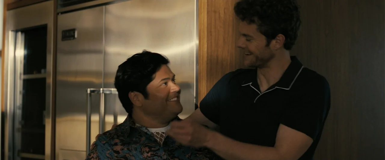 Companion (2025) movie still: medium — A modern home interior with a stainless steel refrigerator and wood paneling on walls. Two men stand facing…
