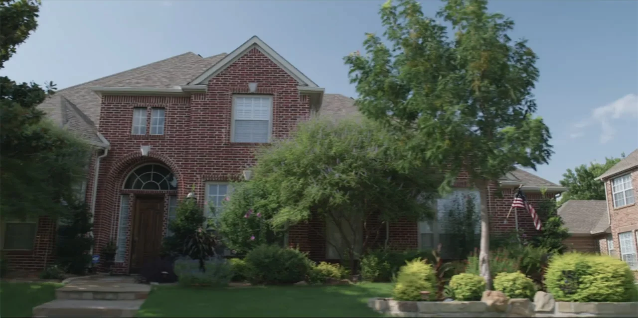 A Radical Life (2022) movie still: establishing — A two-story red brick suburban house stands on a manicured green lawn. A small American flag hangs…