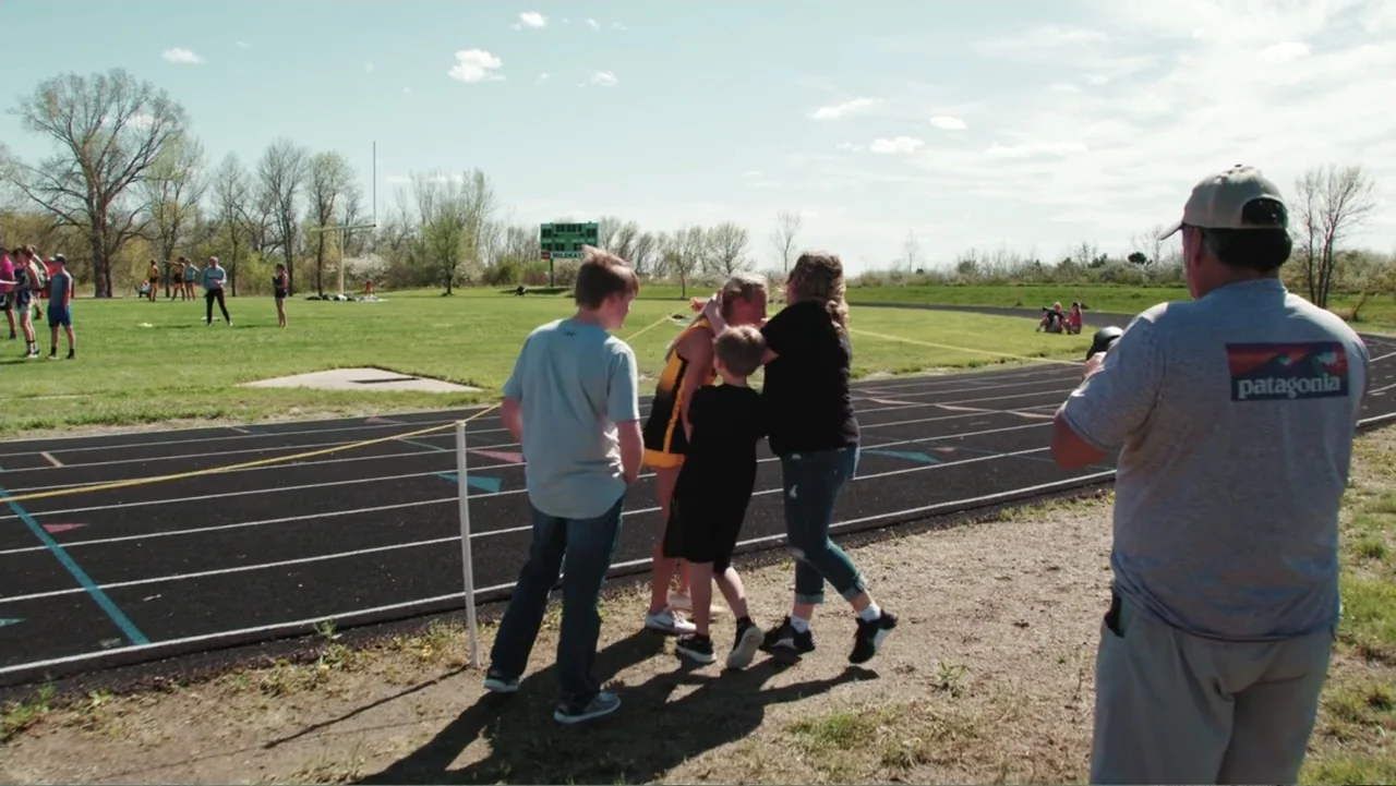 Common Ground (2000) movie still: fullbody — A sunny day at an outdoor running track with grass and trees in background. A group of people gather on…